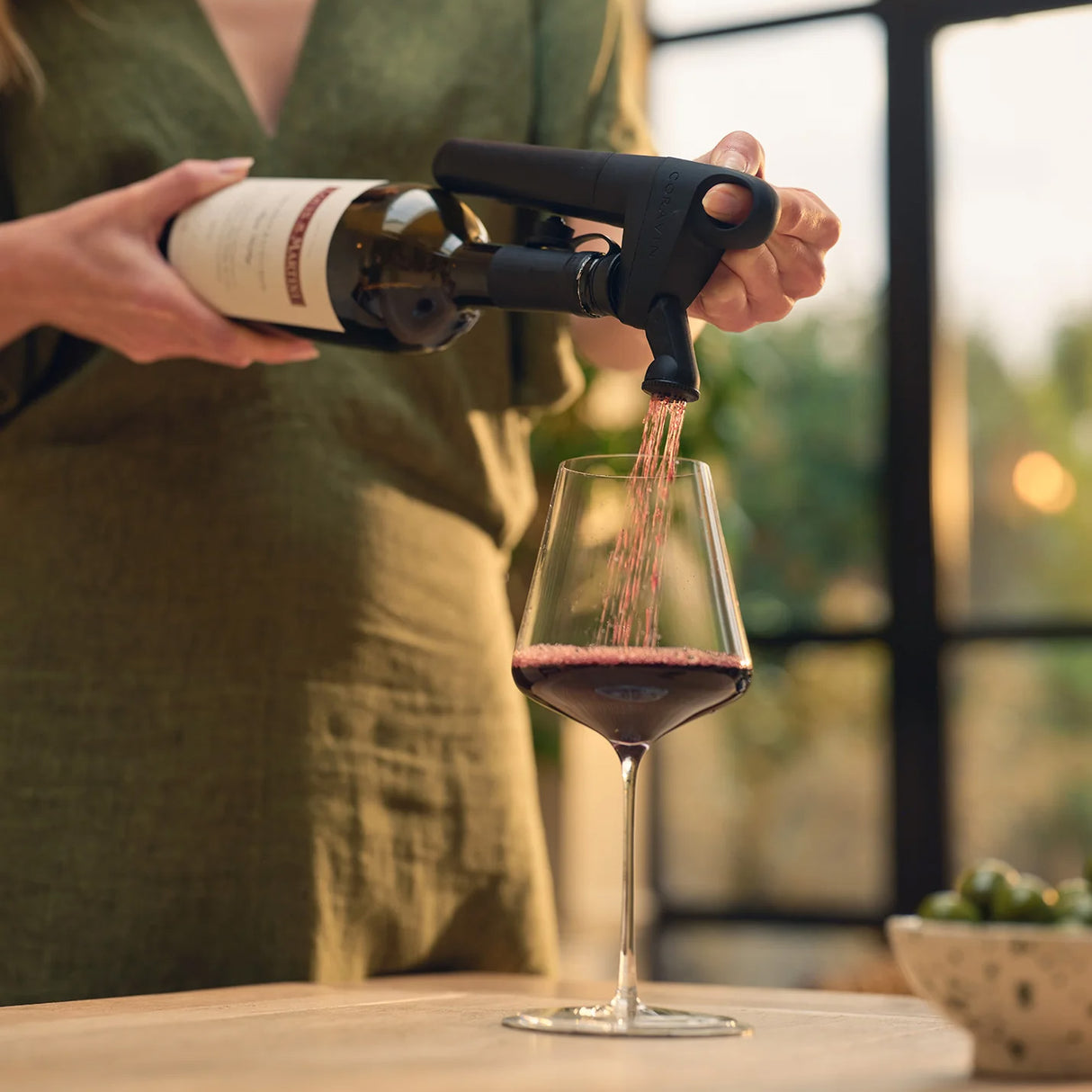 A person in a green dress uses the Coravin Pivot+ Wine Preservation System - Black by Coravin to pour red wine from a bottle into a glass, keeping the wine fresh, while a bowl of olives sits on the nearby table.