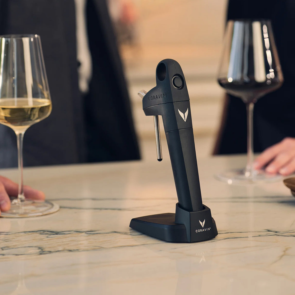 A Coravin Pivot+ Wine Preservation System - Black by Coravin rests on a marble countertop between two people holding glasses of white and red wine. The elegant scene is ideal for a wine tasting, emphasizing the sophistication of this innovative preservation system.