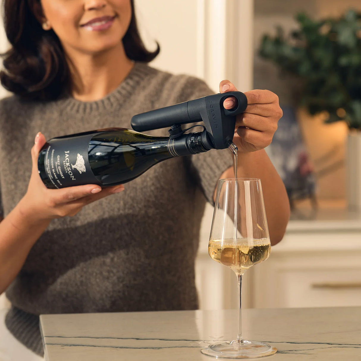 A woman uses the Coravin Pivot+ Wine Preservation System by Coravin in black to pour white wine from a bottle into a glass in a modern kitchen.