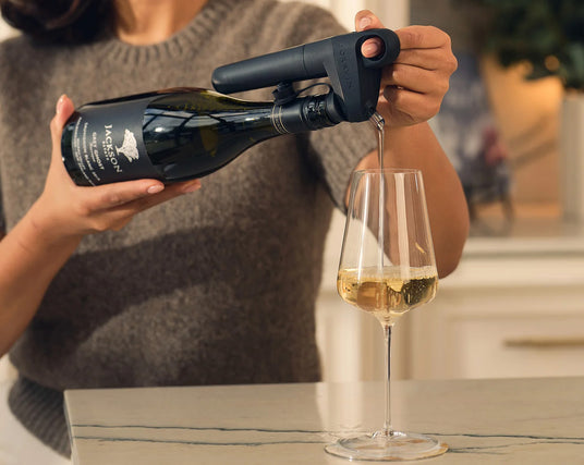 A person in a grey sweater uses a dark gray wine pourer to pour white wine from a bottle into a glass on a kitchen counter.