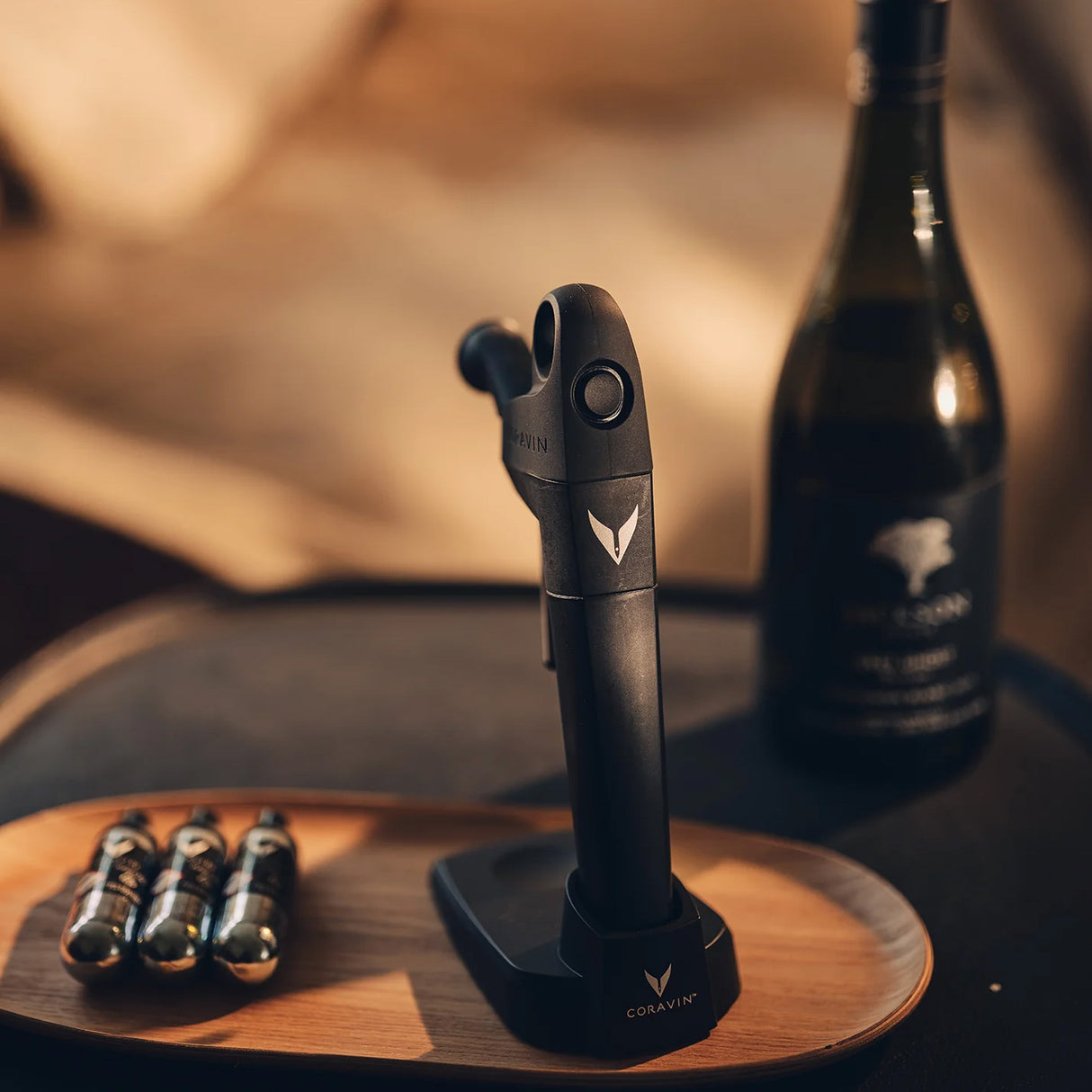 A Coravin Pivot+ Wine Preservation System by Coravin, displayed on a tray with gas capsules beside a wine bottle, illuminated in soft warm lighting.
