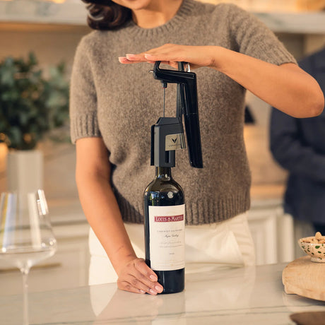 A person in a grey sweater uses a Coravin Timeless Six+ Wine Preservation System in black by Coravin to pour a glass of Louis M. Martini Cabernet Sauvignon. The bottle is placed on a white marble countertop beside an empty wine glass and a small bowl of nuts.