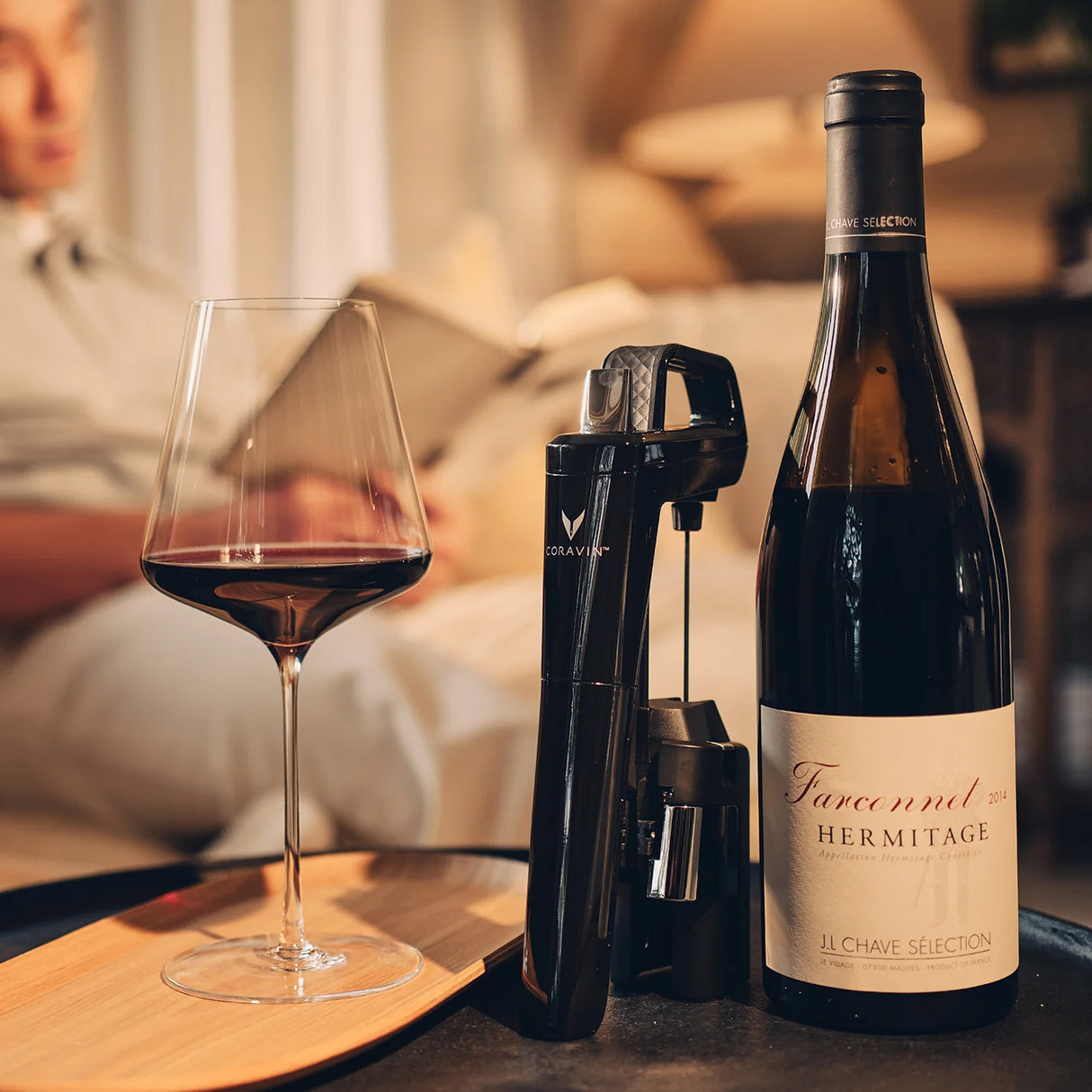 A glass of red wine, a wine opener, and a Coravin Timeless Six+ Wine Preservation System in Black by Coravin are arranged on a table. In the softly blurred background, someone reads a book on a sofa in a cozy living room.