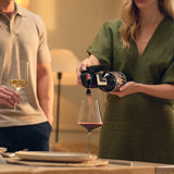 A woman in a green dress uses the Coravin Timeless Six+ Wine Preservation System by Coravin in Black to pour red wine into a glass. Next to her, a man in a beige polo shirt holds a glass of white wine. They are seated in a warm, dimly lit room, likely at a dinner table.