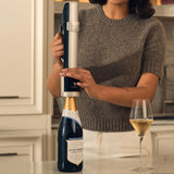 A woman uses the Coravin Sparkling Wine Preservation System by Coravin, featuring a silver and black design, to open a bottle of Nyetimber sparkling wine; beside her on the kitchen counter is a filled wine glass, illustrating how the Coravin system helps maintain freshness and flavor in sparkling wines.