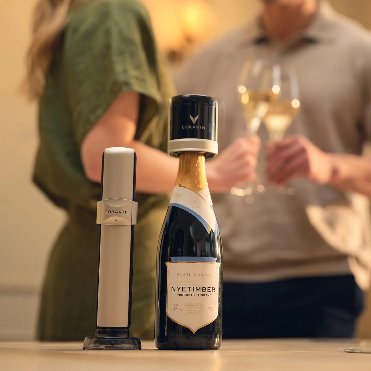 A Coravin Sparkling Wine Preservation System by Coravin sits on a table alongside a bottle of Nyetimber sparkling wine, showcasing an innovative method for preserving sparkling wine with CO2 capsules. In the background, two people holding wine glasses appear out of focus.