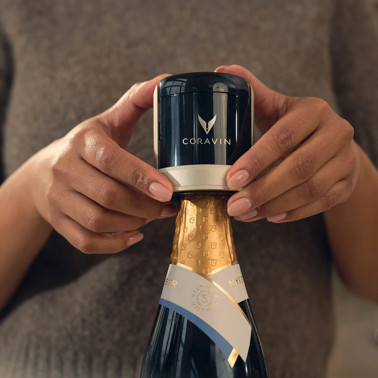 A person in a brown sweater places the Coravin Sparkling Wine Preservation System by Coravin on top of a sealed champagne bottle, demonstrating how the included CO₂ capsules are used to preserve sparkling wine.