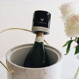A wine bottle with a Coravin Sparkling Wine Stopper by Coravin attached sits in an ice bucket next to a vase of white flowers, elegantly showcasing the innovative set of two stoppers designed specifically for preserving sparkling wine bottles.