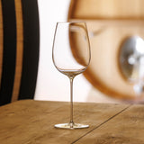 A clear, mouth-blown crystal Sydonios Racine Collection - l’Esthète Red Wine Glass from a set of 6 sits on a wooden table. The softly blurred background reveals part of a wooden barrel and a white wall.