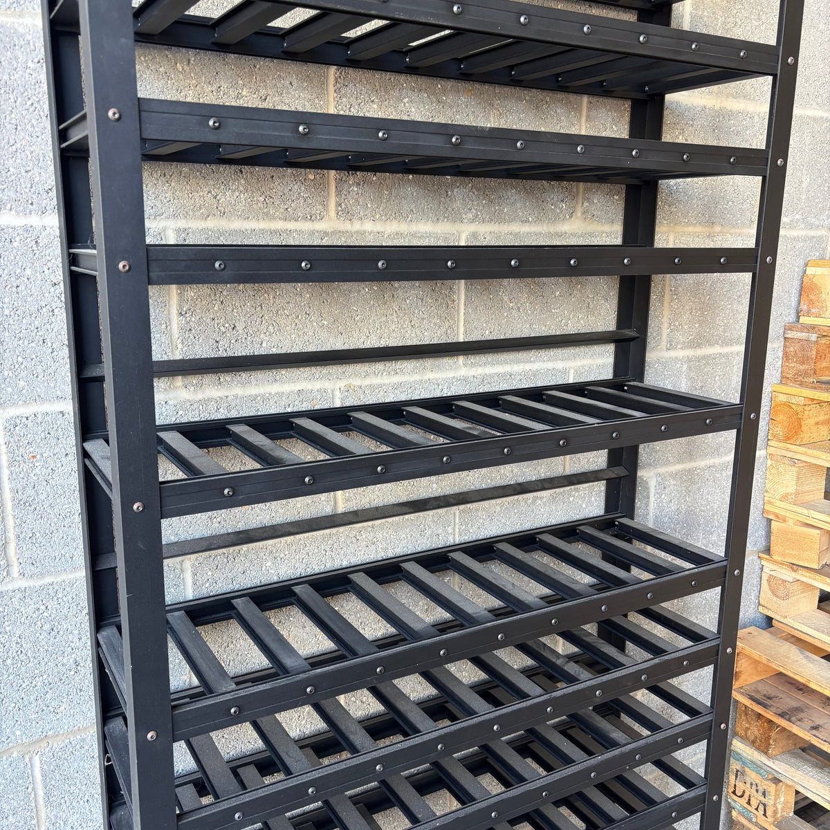 A tall black metal wine rack with multiple slatted shelves, photographed against a gray cinderblock wall with wooden pallets partially visible on the right.