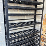 A tall black metal wine rack with multiple slatted shelves, photographed against a gray cinderblock wall with wooden pallets partially visible on the right.