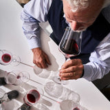 An older man with curly gray hair smells a glass of red wine at a white table set with several partially filled Gabriel-Glas StandArt Universal Red & White Wine Glasses by Gabriel-Glas, from the Set of 2.