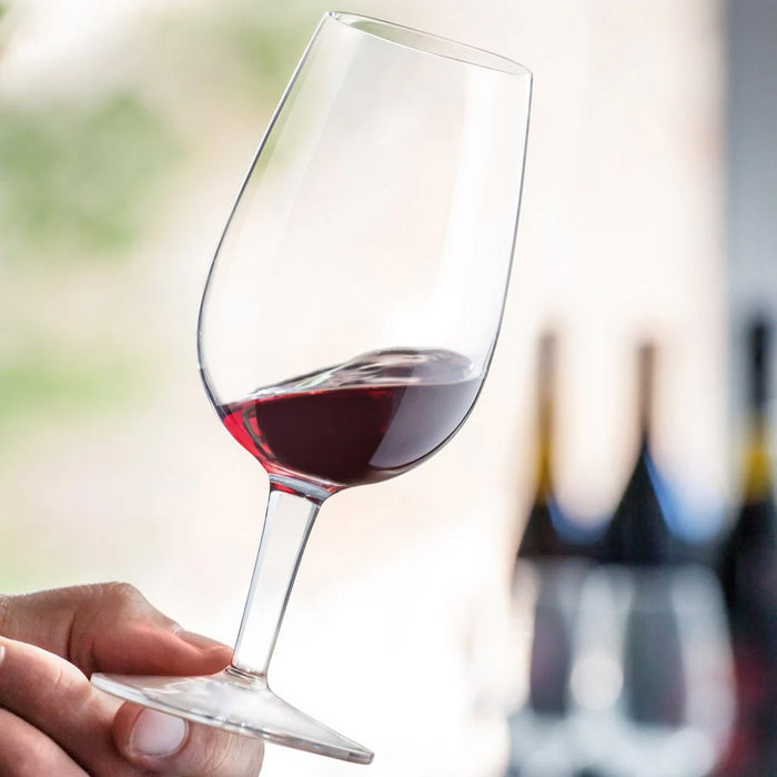 A hand holds a Luigi Bormioli ISO Type Wine Tasting Glass (21.5cl) at an angle, with blurred wine bottles behind—ideal for professional tastings or advancing your wine knowledge. Set of 6 glasses by Luigi Bormioli.