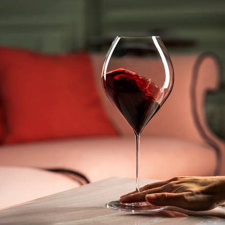 A hand rests on a table beside a mouth-blown Lehmann Glass La Poétique Grand Rouge Red Wine Glass 700ml filled with swirling red wine.