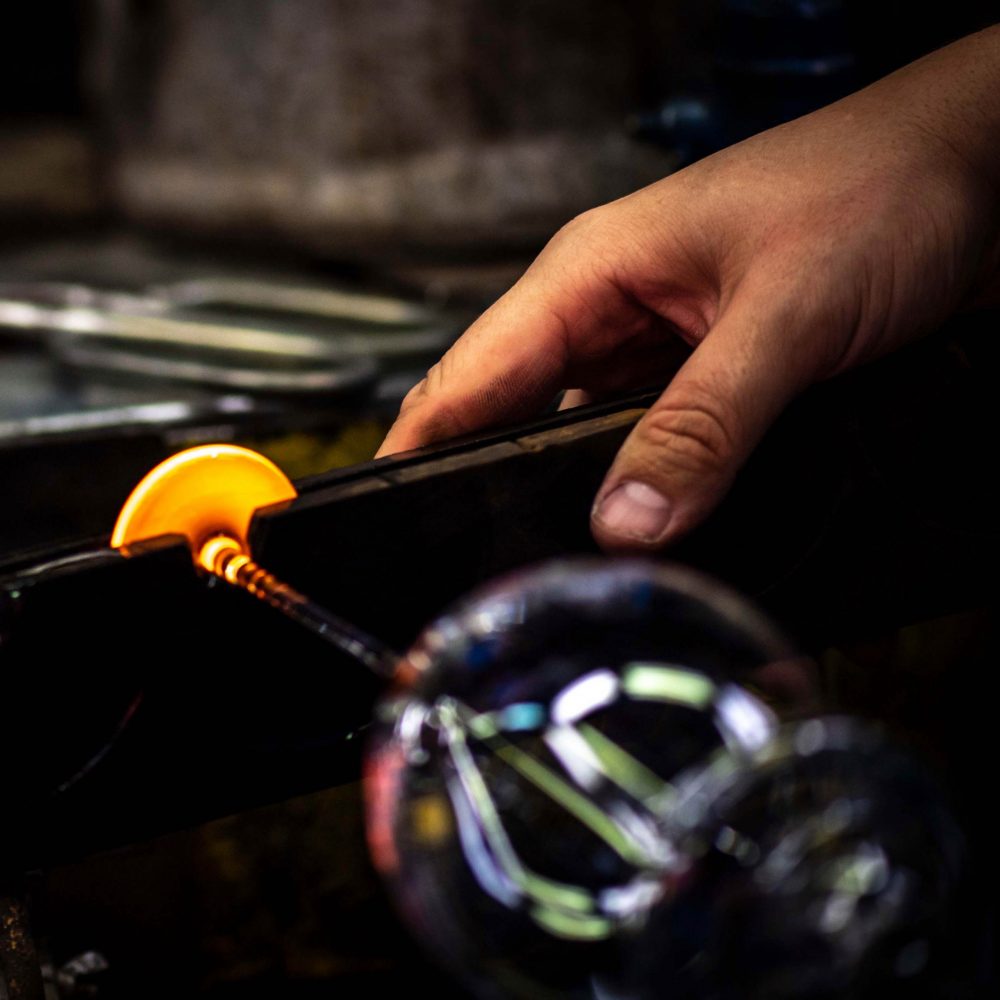 A glassblower’s hand uses a tool to shape the glowing orange base of a mouth-blown Lehmann Glass La Poétique Grand Rouge Red Wine Glass 700ml.