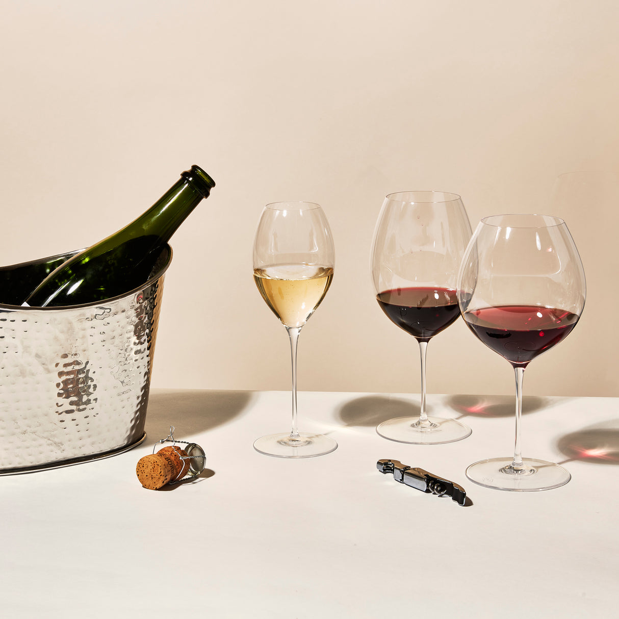 A bottle of wine sits in a silver ice bucket beside two Richard Brendon The Occasional Collection Champagne/Sparkling Wine Glasses. Nearby, a cork, wire cage, and corkscrew rest on a white surface.