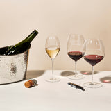 A bottle of wine sits in a silver ice bucket beside two Richard Brendon The Occasional Collection Champagne/Sparkling Wine Glasses. Nearby, a cork, wire cage, and corkscrew rest on a white surface.
