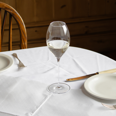 A Jancis Robinson x Richard Brendon The Occasional Collection Champagne/Sparkling Wine Glass - Set of 2 filled with white wine sits on a round table with a white tablecloth, two plates, cutlery, beside a wooden chair and paneled wall.