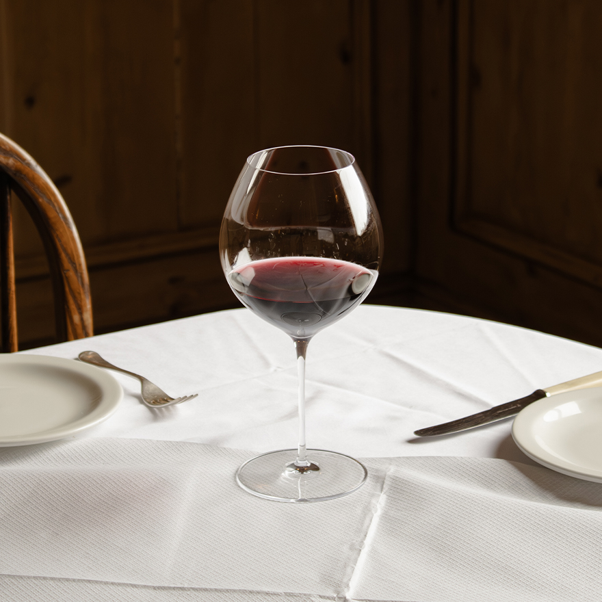 A set of two Richard Brendon The Occasional Collection Burgundy Wine Glasses by Jancis Robinson x Richard Brendon rests on a white tablecloth between plates and cutlery, evoking an inviting wood-paneled dining atmosphere for two.