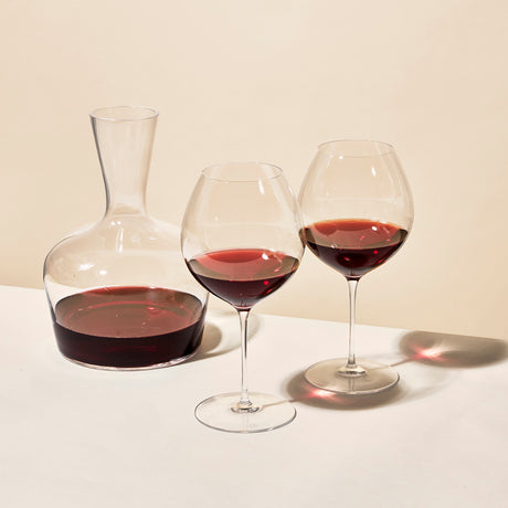 A mouth-blown crystal decanter of red wine sits beside a Richard Brendon The Occasional Collection Burgundy Wine Glass - Set of 2, each glass partially filled, on a white surface with a neutral beige background.