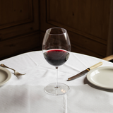 A set of two Jancis Robinson x Richard Brendon The Occasional Collection Bordeaux Wine Glasses rests on a white tablecloth between plates with cutlery, their mouth-blown crystal bringing elegance to the warmly-lit dining scene.