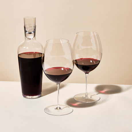A clear glass decanter with red wine sits next to two half-filled Richard Brendon The Occasional Collection Bordeaux Wine Glasses (Set of 2) on a white surface, casting soft shadows against a beige background.