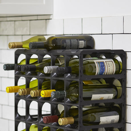 A pair of Stakrax Plastic Wine Rack Black Top Plates by Stakrax holds multiple wine bottles horizontally against a white tiled kitchen wall, with some bottles extending out from the modular rack.