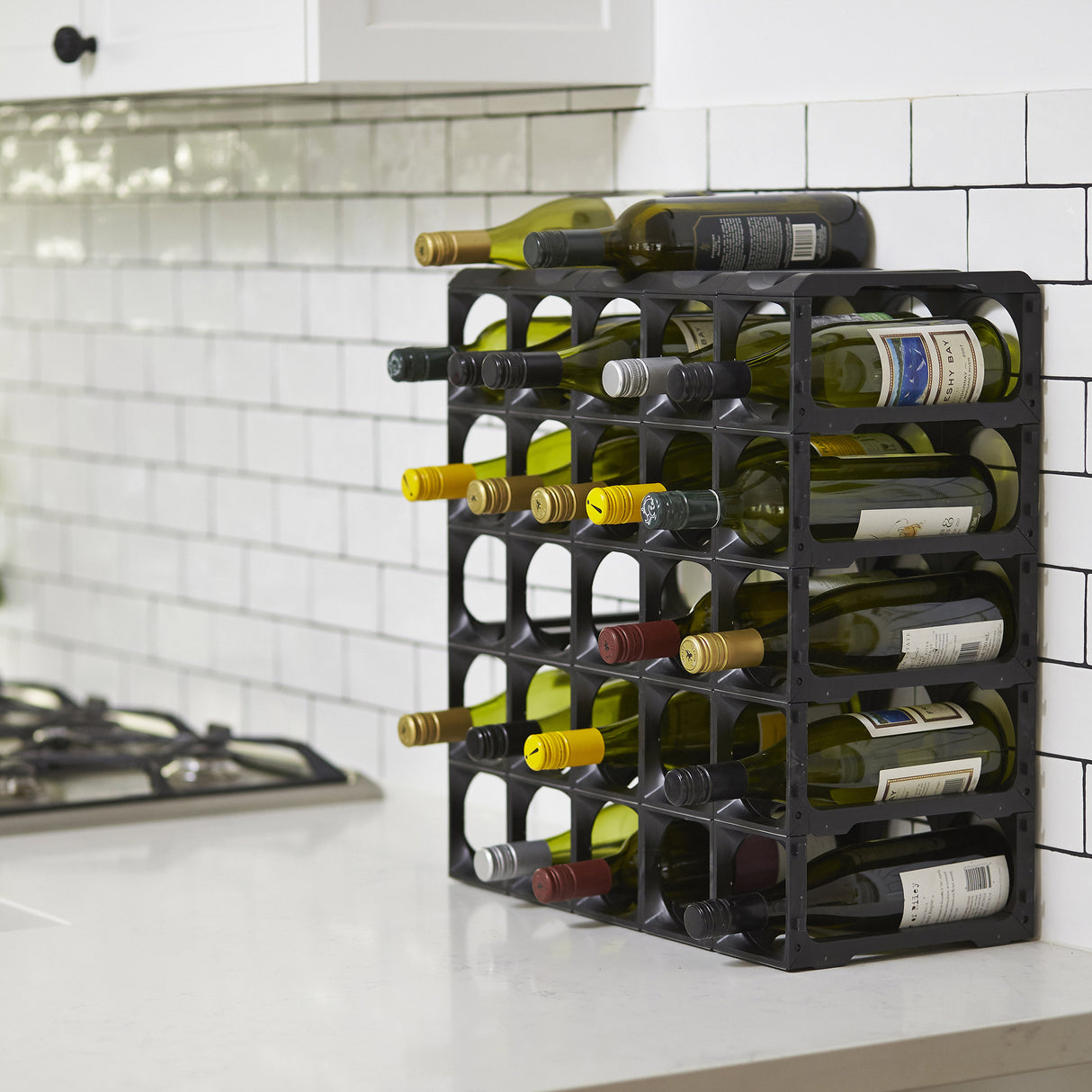 The Stakrax 30 Bottle Plastic Wine Rack in black by Stakrax, filled with wine bottles, sits on a white kitchen counter against white subway tile, next to a stainless steel gas stove.
