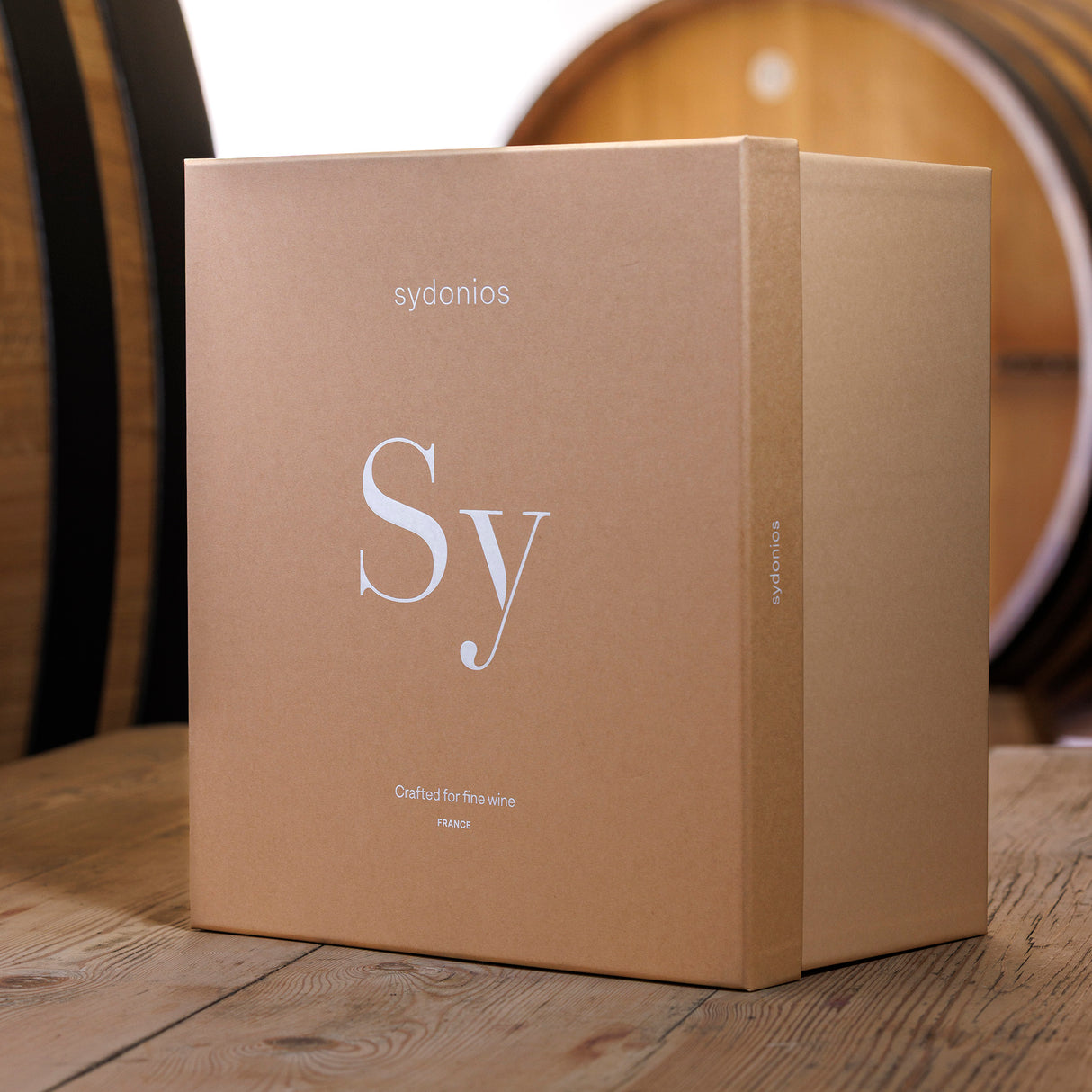 A large, light brown Sydonios box marked with "Sy" sits on a wooden table, hinting at the exquisite Sydonios Racine Collection - l’Esthète Red Wine Glasses (Set of 2) inside. Blurred wine barrels fill the background.