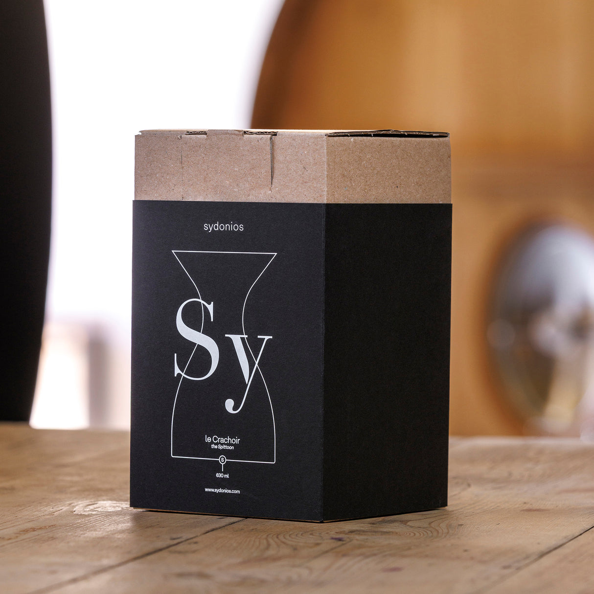 A rectangular black and brown cardboard box labeled Sydonios rests on a wooden table. Minimalist white graphics suggest it contains the Sydonios Small Wine Tasting Spittoon 630ml – Black, designed for professional wine tastings.
