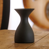 A matte black, hourglass-shaped object—the Sydonios Small Wine Tasting Spittoon 630ml by Sydonios—rests on a wooden table against a blurred background of warm tones and rounded shapes. This professional wine tasting accessory, crafted from mouth-blown glass, combines elegance with practical functionality.