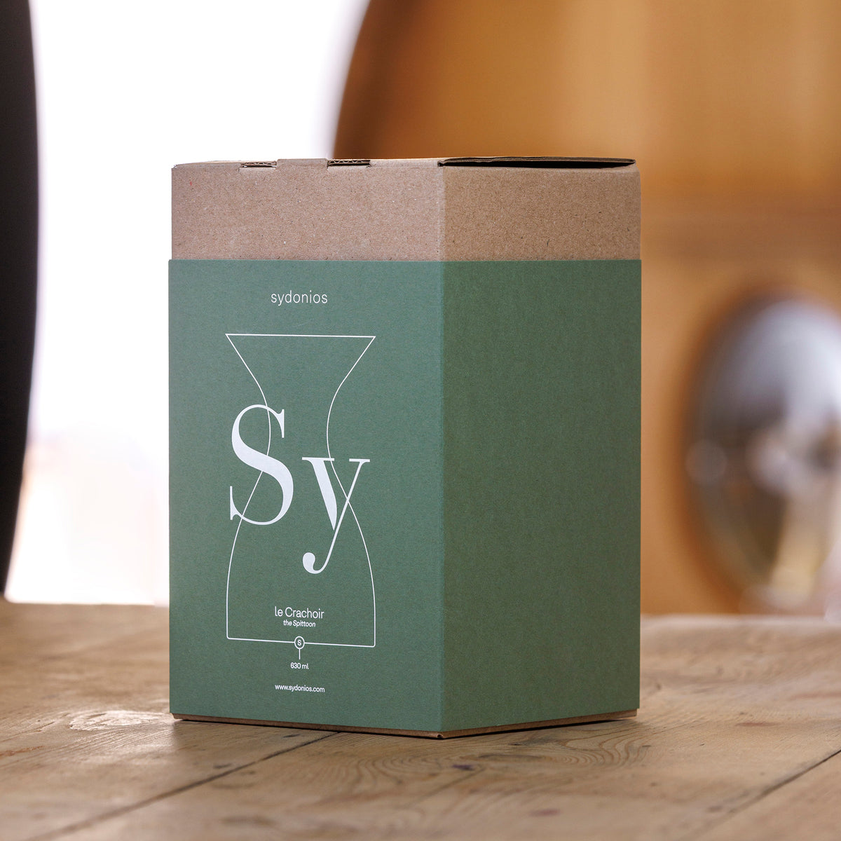 A green and brown cardboard box labeled with the Sydonios logo rests on a wooden surface, featuring a minimalist outline of a carafe and the names “Sydonios” and “Le Crachoir” on the front. This packaging hints at the refined design of the Sydonios Small Wine Tasting Spittoon 630ml - Jade (Green), a mouth-blown glass spittoon commonly used in professional wine tastings. The background is softly blurred.