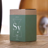 A green and brown cardboard box labeled with the Sydonios logo rests on a wooden surface, featuring a minimalist outline of a carafe and the names “Sydonios” and “Le Crachoir” on the front. This packaging hints at the refined design of the Sydonios Small Wine Tasting Spittoon 630ml - Jade (Green), a mouth-blown glass spittoon commonly used in professional wine tastings. The background is softly blurred.