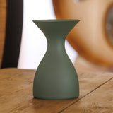 A Sydonios Small Wine Tasting Spittoon 630ml in Jade (Green), featuring a matte, mouth-blown glass hourglass shape, sits on a wooden table with a blurred, warmly lit background.