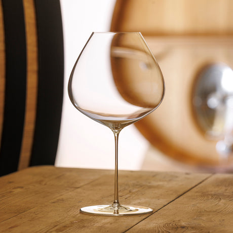 A set of six Sydonios Terroir Collection Le Septentrional red wine glasses with wide bowls rests on a wooden table, a softly blurred wine barrel in the background—ideal for enjoying matured red wines.