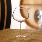 A set of six Sydonios Racine Collection le Subtil red wine glasses sits on a wooden table, their wide, mouth-blown crystal bowls inviting appreciation of fine wines. Wooden barrels in the warmly lit background evoke oenological precision.