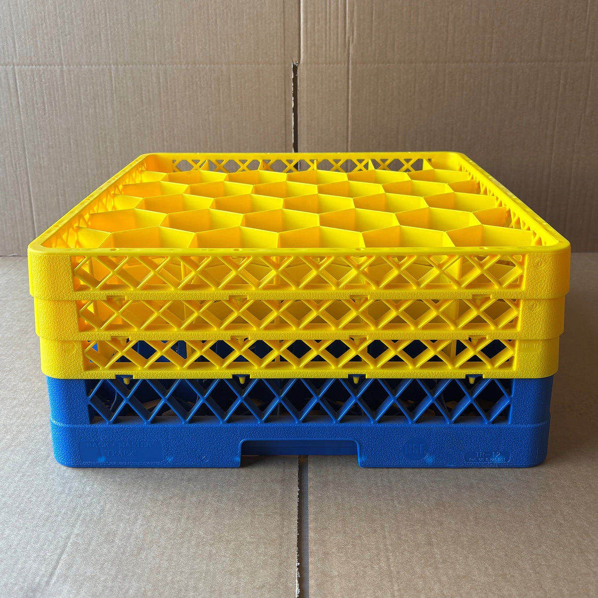 A stack of three yellow and one blue Wineware Traex Rack Master Glass Washer Trays (USED - GREAT CONDITION, 500 x 500 x 200mm, holds 30 glasses, 81mm cell) sits on a cardboard surface with a cardboard background.