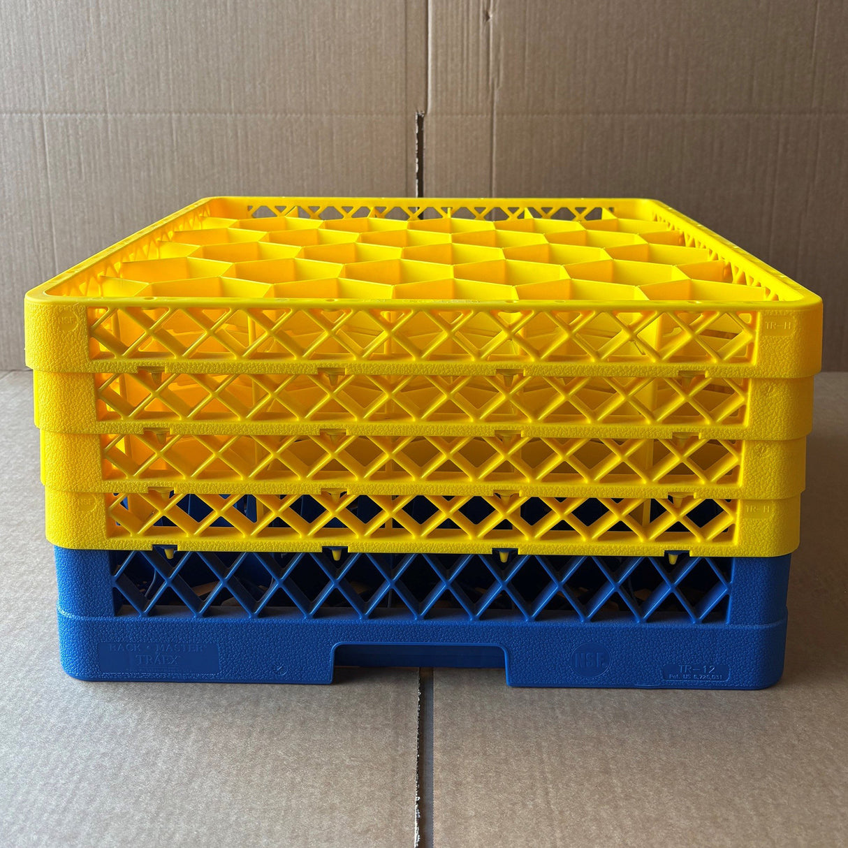 A stack of four Wineware Traex Rack Master Glass Washer Trays (USED - GREAT CONDITION, 500x500x240mm, holds 30 glasses with 81mm cells), three yellow and one blue, is placed on a cardboard surface in front of a cardboard background.