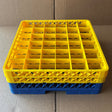 A Wineware Traex Rack Master Glass Washer Tray (500x500x160mm, 36 glasses, 75mm cell) in yellow plastic is stacked on a blue rack, both resting on cardboard.