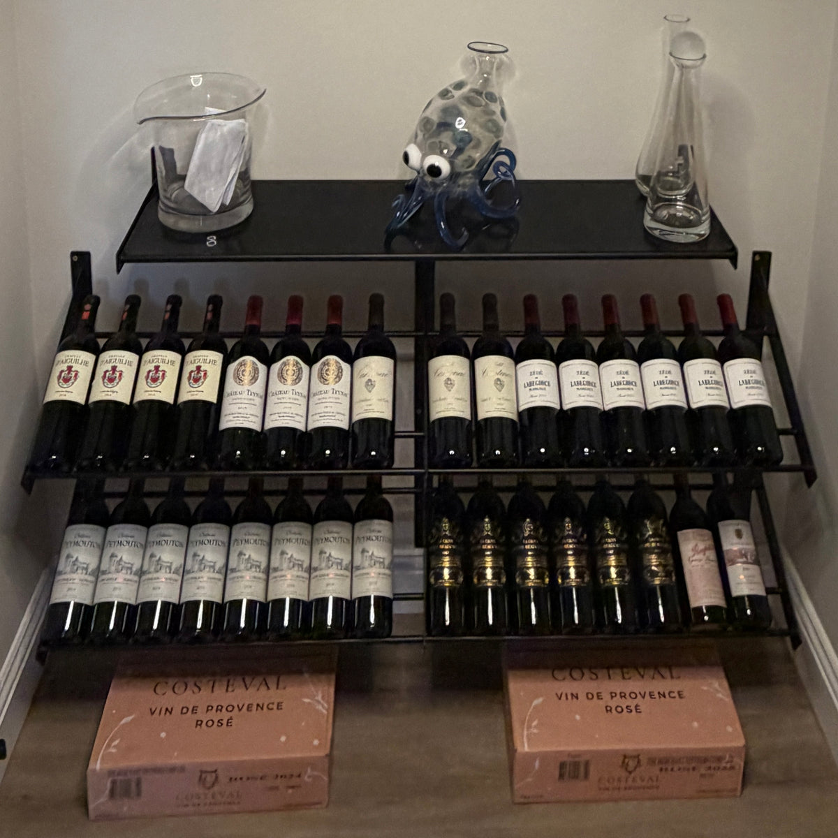 A VintageView Wall Mounted W Series Presentation Wine Rack by VintageView holds three rows of red wine bottles, with glass decanters, a glass fish, napkins, and two boxes of rosé wine arranged on the floor beneath. This elegant wine bottle display is showcased in the corner of the room.