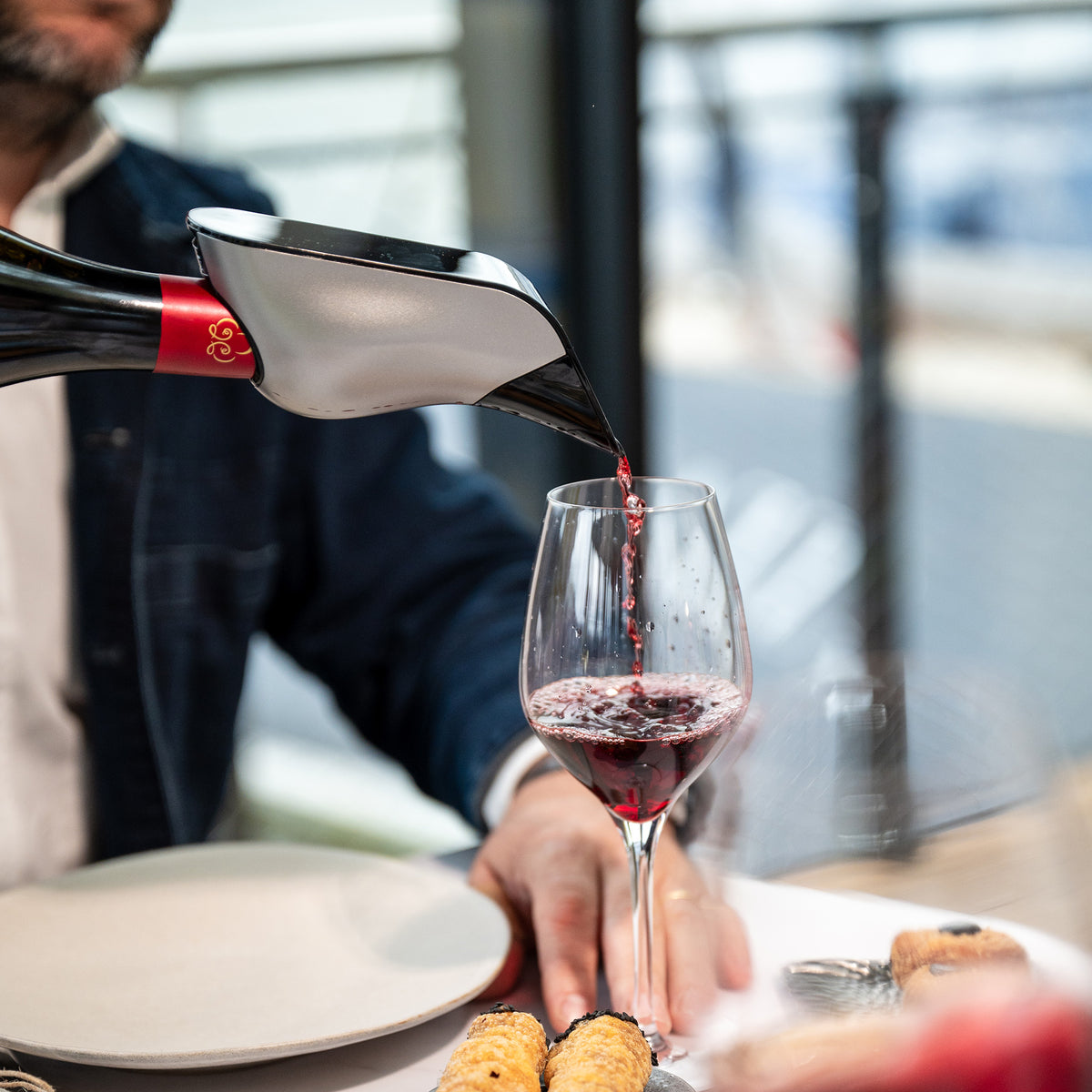 A person uses the Aveine Original Smart Wine Aerator by Aveine to pour red wine from a decanter into a wine glass at a table set with plates and pastries, instantly providing 1–24 hours of aeration for enhanced aroma and flavor.