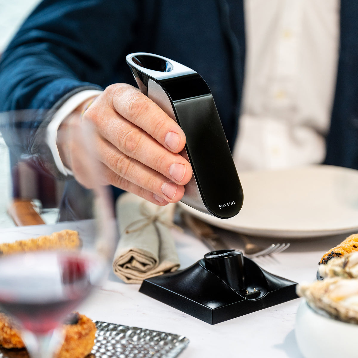 A person in a suit holds the Aveine Original Smart Wine Aerator by Aveine—an elegant black device that delivers instant 1–24 hour aeration—over a plate at a dining table set with food, wine, and a napkin, highlighting how advanced aeration technology enhances the dining experience.