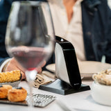 The Aveine Original Smart Wine Aerator by Aveine, a sleek and modern device, sits on a dining table alongside a glass of red wine, food, and a person in a suit blurred in the background, effortlessly delivering instant 1–24 hour aeration to every pour with advanced micro-oxygenation technology.