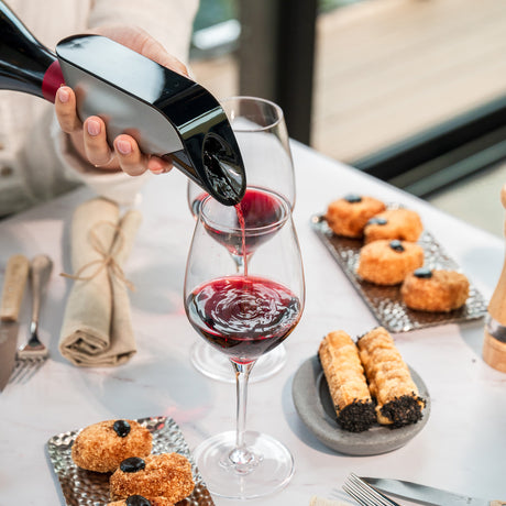 At a table set with pastries and rolled wafer cookies, next to a napkin, utensils, and a bright window, someone pours red wine into a glass using the Aveine Original Smart Wine Aerator by Aveine, instantly enhancing the flavor with advanced wine aeration for 1 to 24 hours.