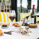 A table set with wine glasses, a bottle of red wine complemented by the Aveine Original Smart Wine Aerator from Aveine for instant 1–24 hour aeration, plates of breaded appetizers, utensils, and napkins, with yellow and white striped seating in the background.
