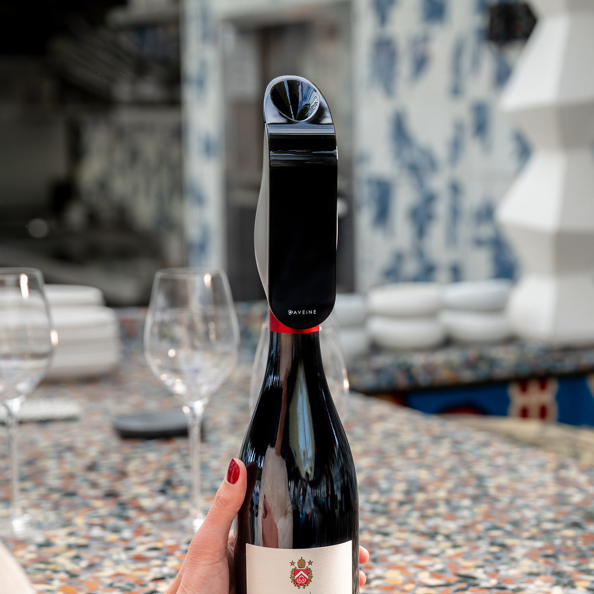 A hand with red nail polish holds a wine bottle fitted with the Aveine Original Smart Wine Aerator by Aveine, delivering instant 1–24 hour aeration through advanced micro-oxygenation technology. In the background, two empty wine glasses and stacked plates sit on a colorful, patterned table.
