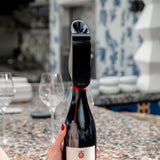 A hand with red nail polish holds a wine bottle fitted with the Aveine Original Smart Wine Aerator by Aveine, delivering instant 1–24 hour aeration through advanced micro-oxygenation technology. In the background, two empty wine glasses and stacked plates sit on a colorful, patterned table.
