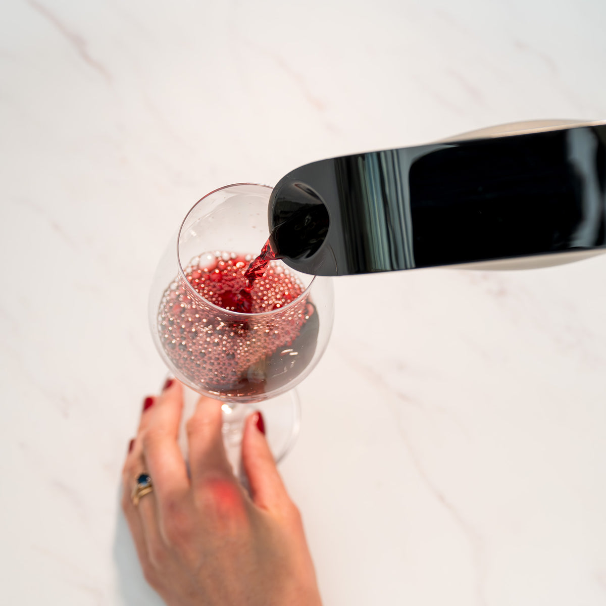 A hand with red-painted nails holds a wine glass while red wine, instantly aerated for optimal micro-oxygenation by the Aveine Original Smart Wine Aerator from Aveine, is being poured into it from a black bottle against a white marble surface.