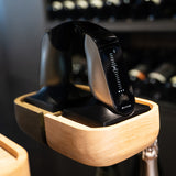 An Aveine Original Smart Wine Aerator by Aveine, featuring a sleek black electronic design and a digital display, sits in a light wood charging dock, its reflection visible on the glass surface with blurred wine bottles in the background—a sophisticated device offering instant 1–24 hour aeration to elevate your wine experience.