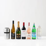 Six bottles—including wine, champagne, rosé, sparkling water, and a soda maker bottle—are lined up in a row with the Huski Wine & Champagne Bottle Cooler by Huski in brushed stainless steel positioned on the left, all set against a white background.